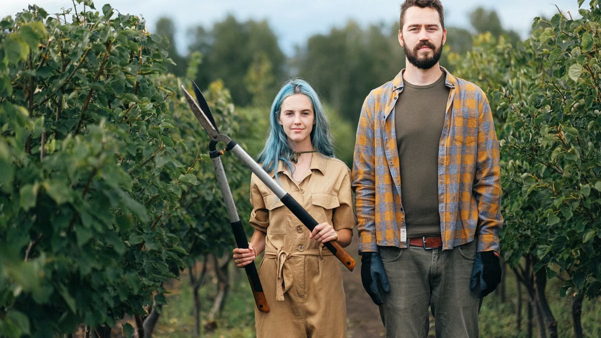A young couple stands in a lush orchard, one holding gardening shears, symbolizing teamwork and organic farming.